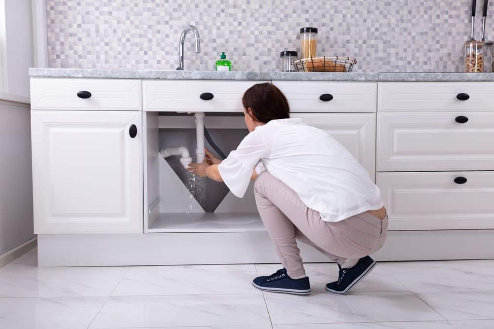 How to Fix a Leaky Pipe Until the Plumber Arrives A woman kneeling down in front of an open kitchen sink.